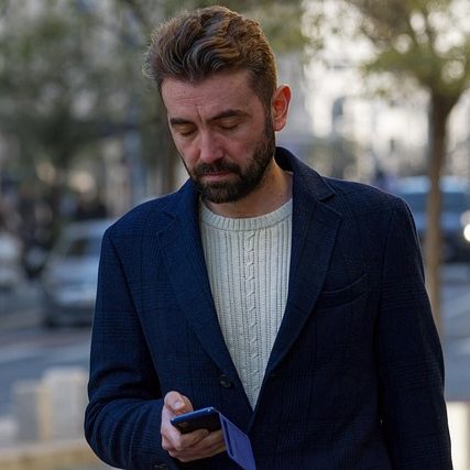 Mann mit Bart und Pullover schaut auf sein Handy, während er draußen läuft.